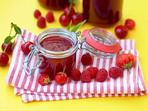 Zwei Einmachgläser mit roter Beerenmarmelade auf gestreiftem Tuch, umgeben von Kirschen, Himbeeren und Erdbeeren auf gelbem Untergrund.
