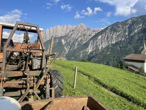 Alter rostiger Traktor auf einer Almwiese mit beeindruckender Alpenkulisse, grünen Hängen und einem Dorf im Hintergrund.