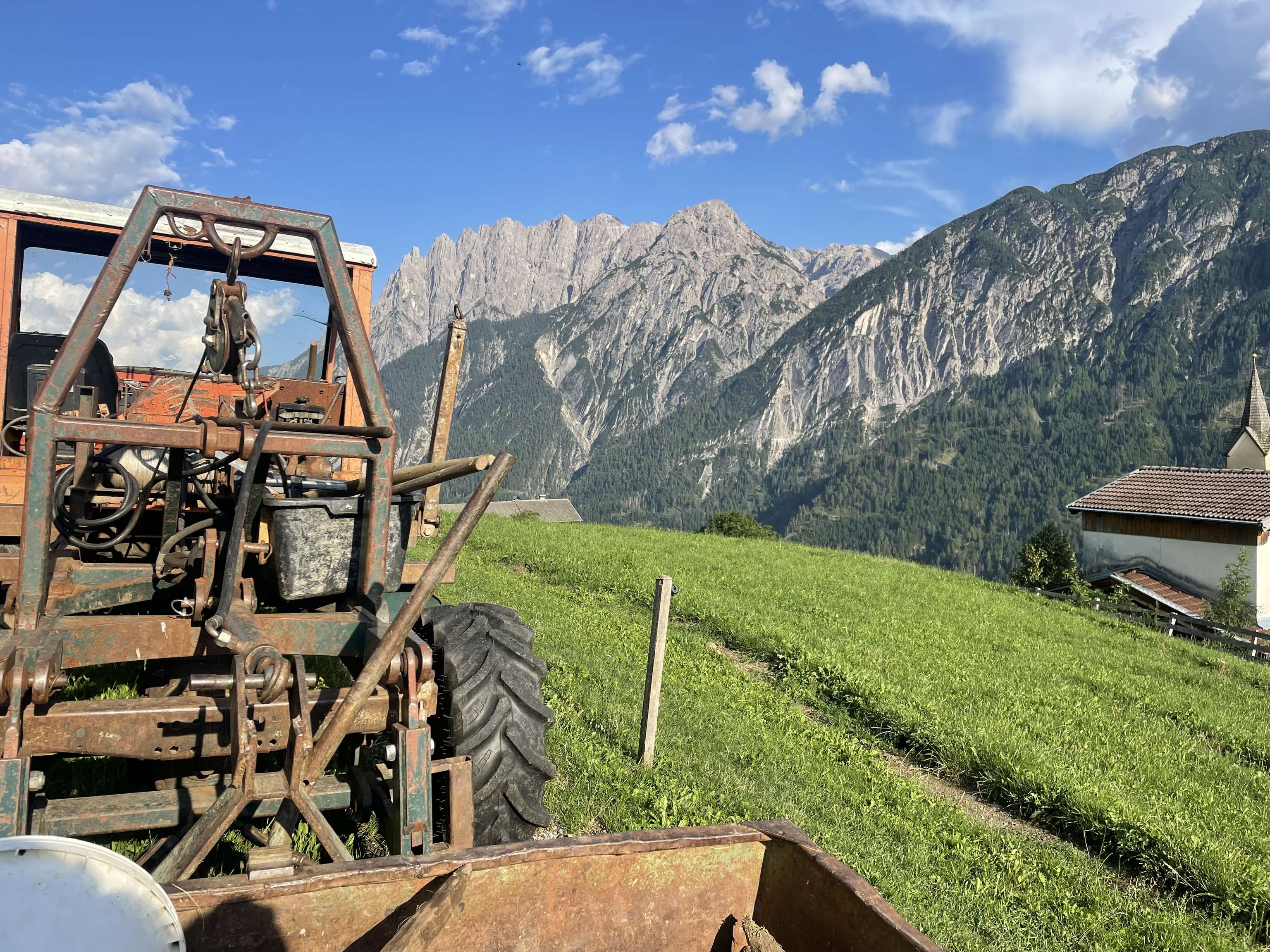 Alter rostiger Traktor auf einer Almwiese mit beeindruckender Alpenkulisse, grünen Hängen und einem Dorf im Hintergrund.
