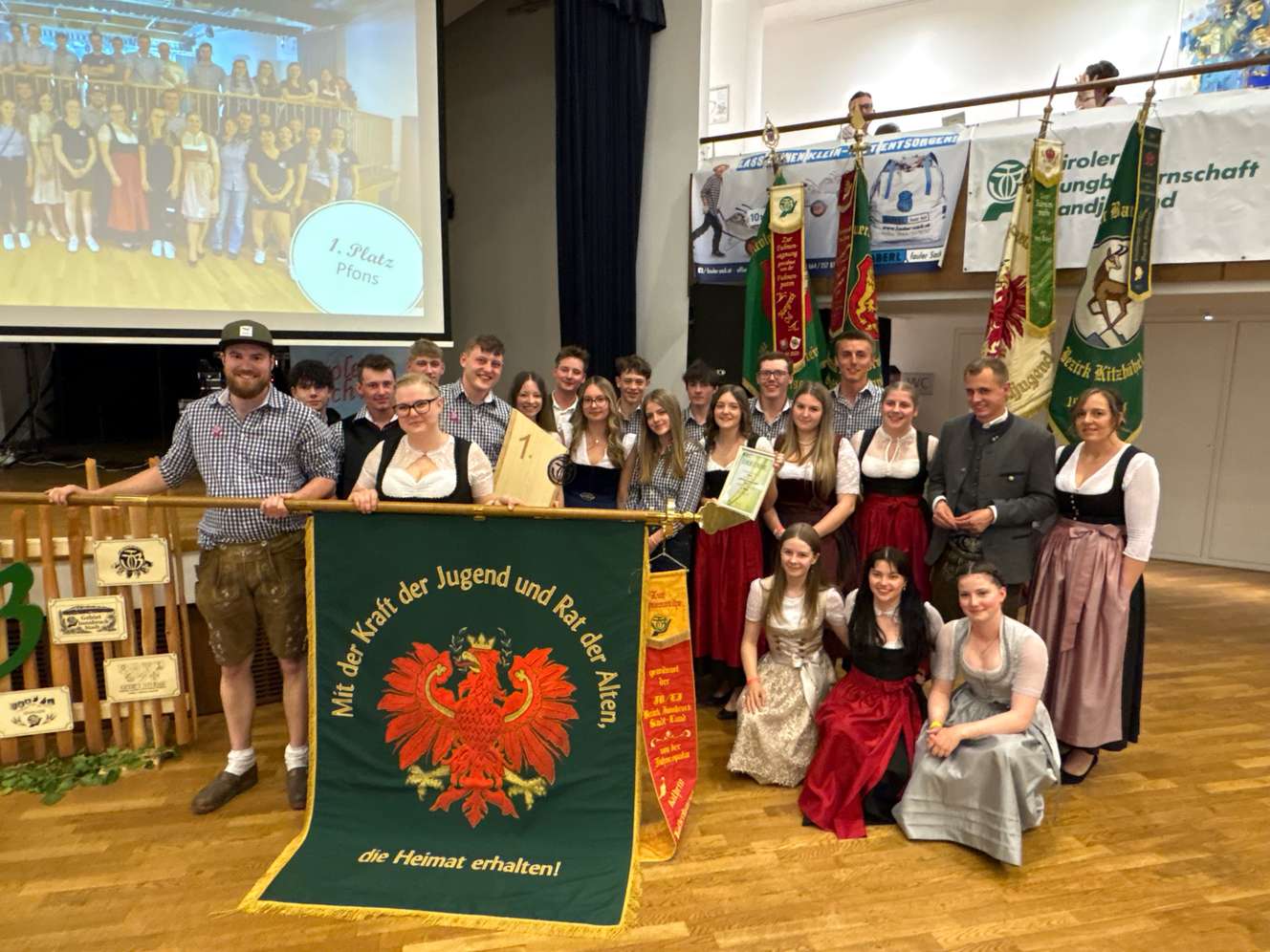Gruppe in traditioneller Tracht posiert mit Banner und Trophäe bei Veranstaltung, im Hintergrund schwenken Fahnen.