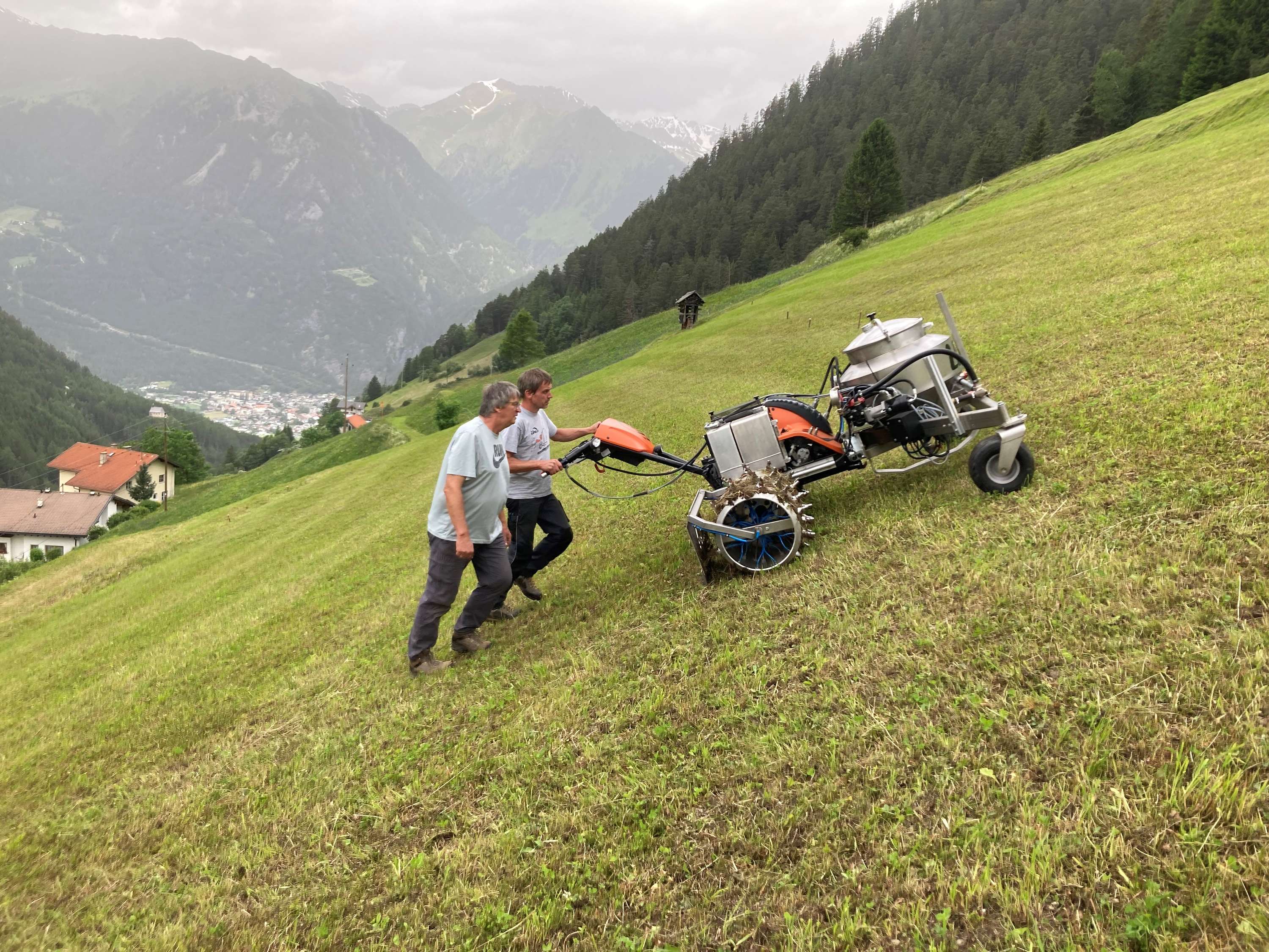Zwei Männer bedienen eine Maschine auf einer steilen Wiese in bergiger Landschaft mit bewölktem Himmel im Hintergrund.