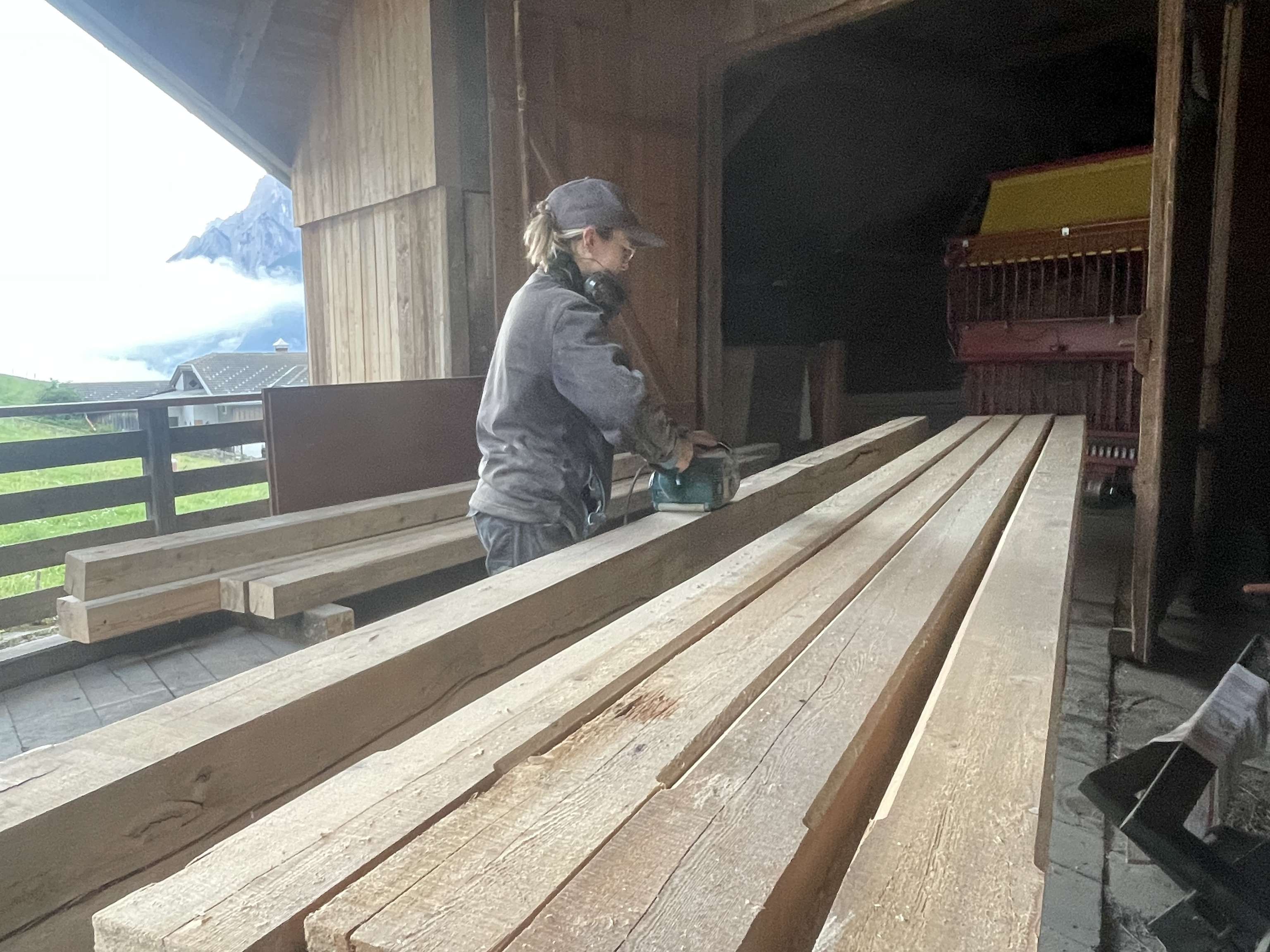 Person schleift lange Holzbalken mit einer Handschleifmaschine auf einer Terrasse mit Bergpanorama im Hintergrund.