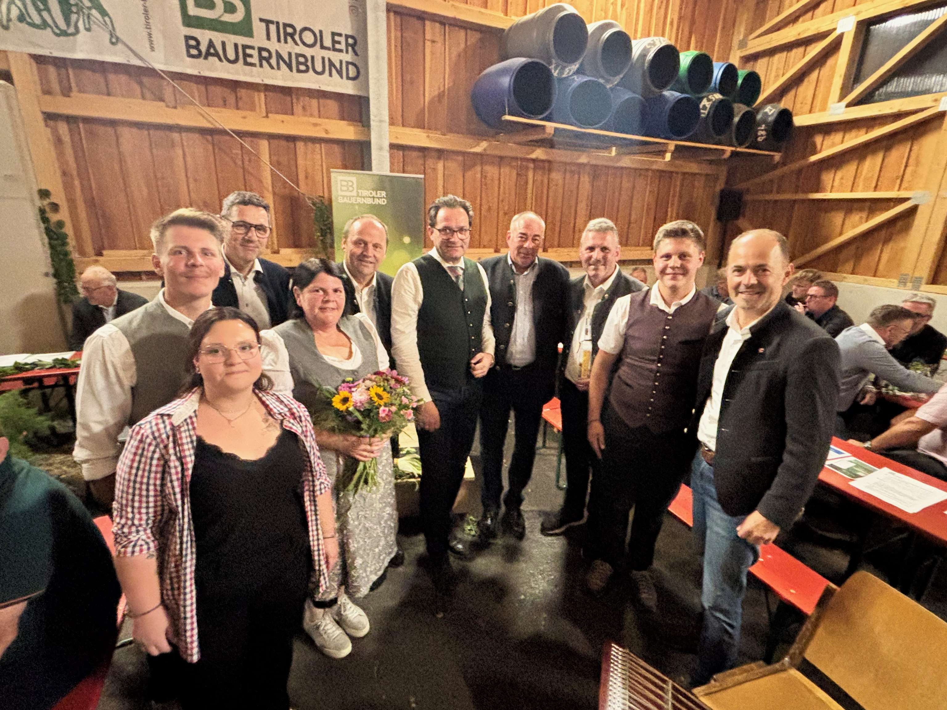 Gruppenfoto beim Tiroler Bauernbund-Event in einer Holzhalle, Frau mit Blumenstrauß im Mittelpunkt, Männer in Tracht.