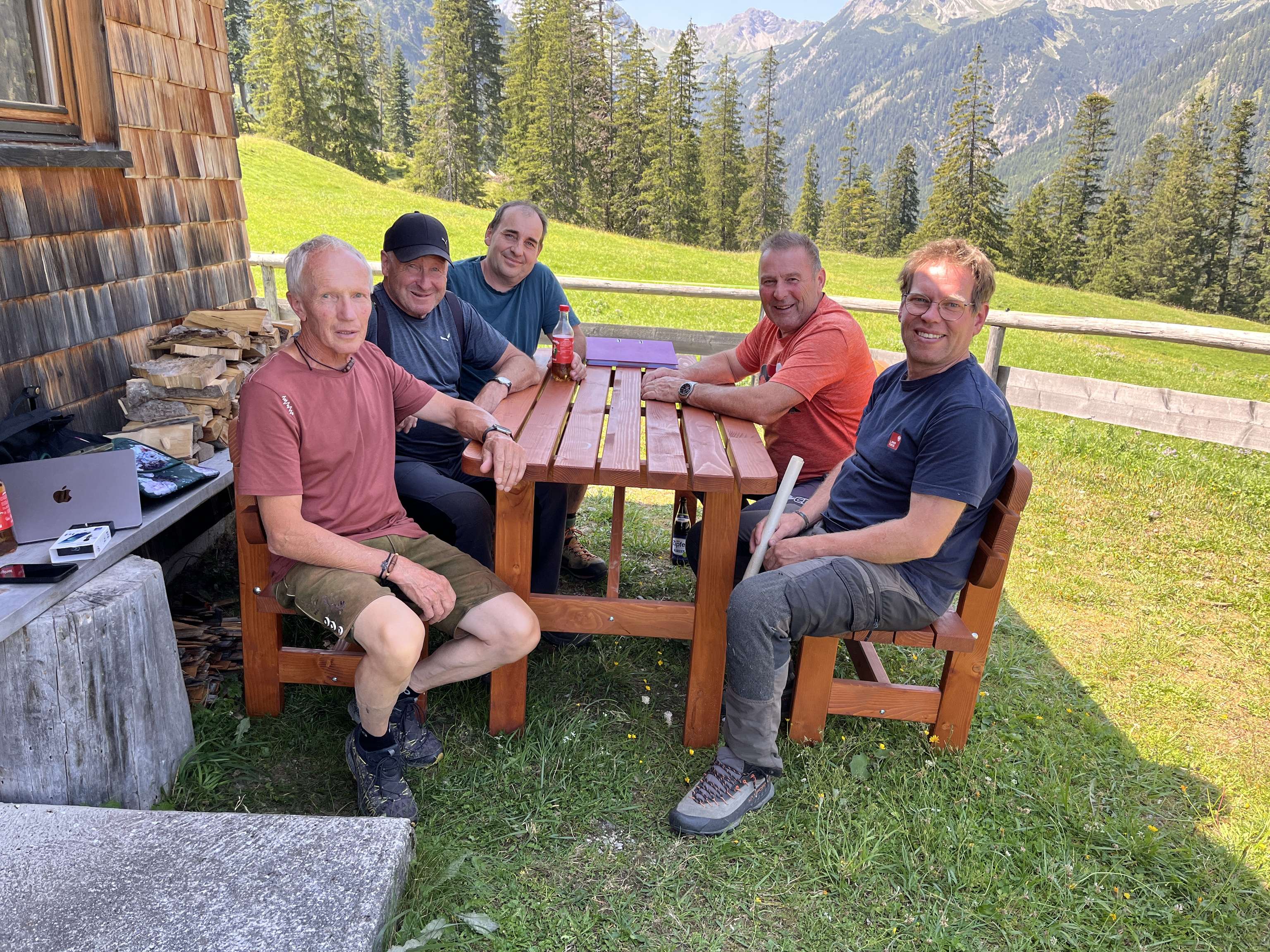 Fünf Männer sitzen an einem Holztisch vor einer Almhütte mit Bergpanorama und Nadelwäldern im Hintergrund.