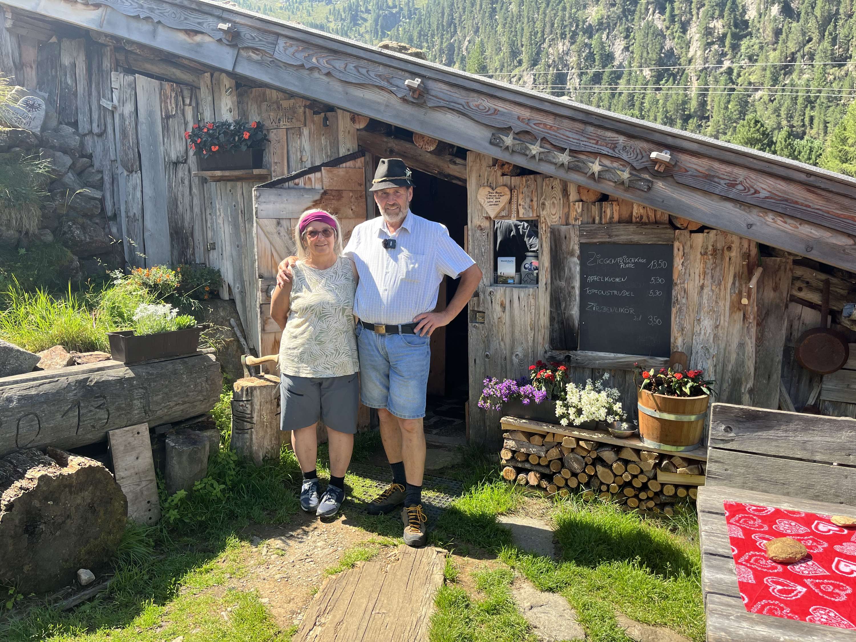 Ruth und Georg Mair stehen lächelnd vor der Almhütte der Mischbachalm in den Bergen mit Speisekarte, Blumen und Holzstapel.