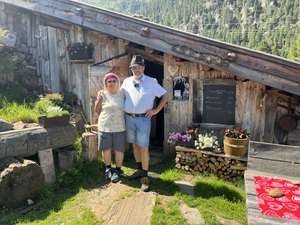 Ruth und Georg Mair stehen lächelnd vor der Almhütte der Mischbachalm in den Bergen mit Speisekarte, Blumen und Holzstapel.