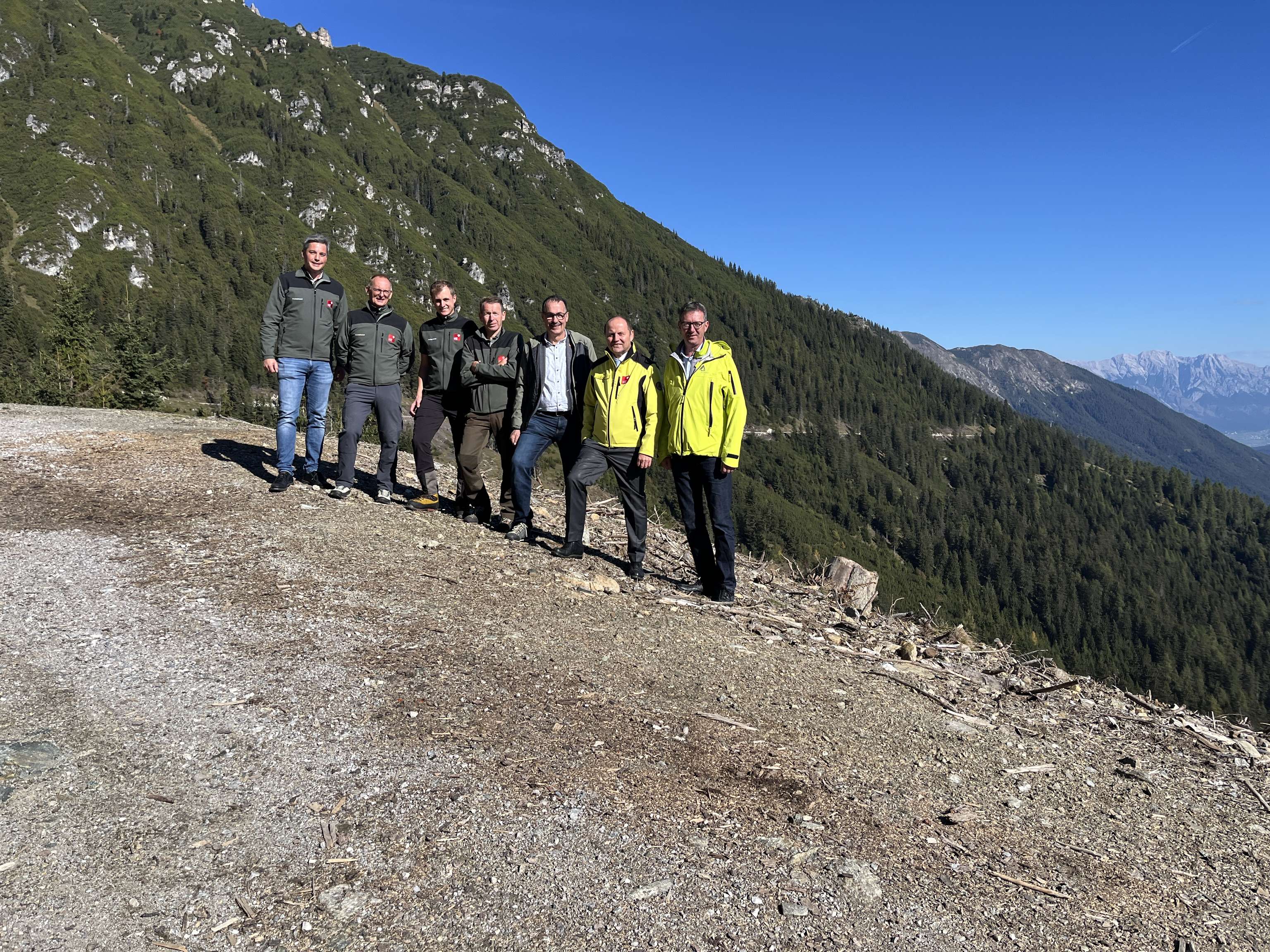 Sieben Männer stehen auf einem Schotterweg in den Alpen mit Blick auf bewaldete Berge und blauem Himmel.