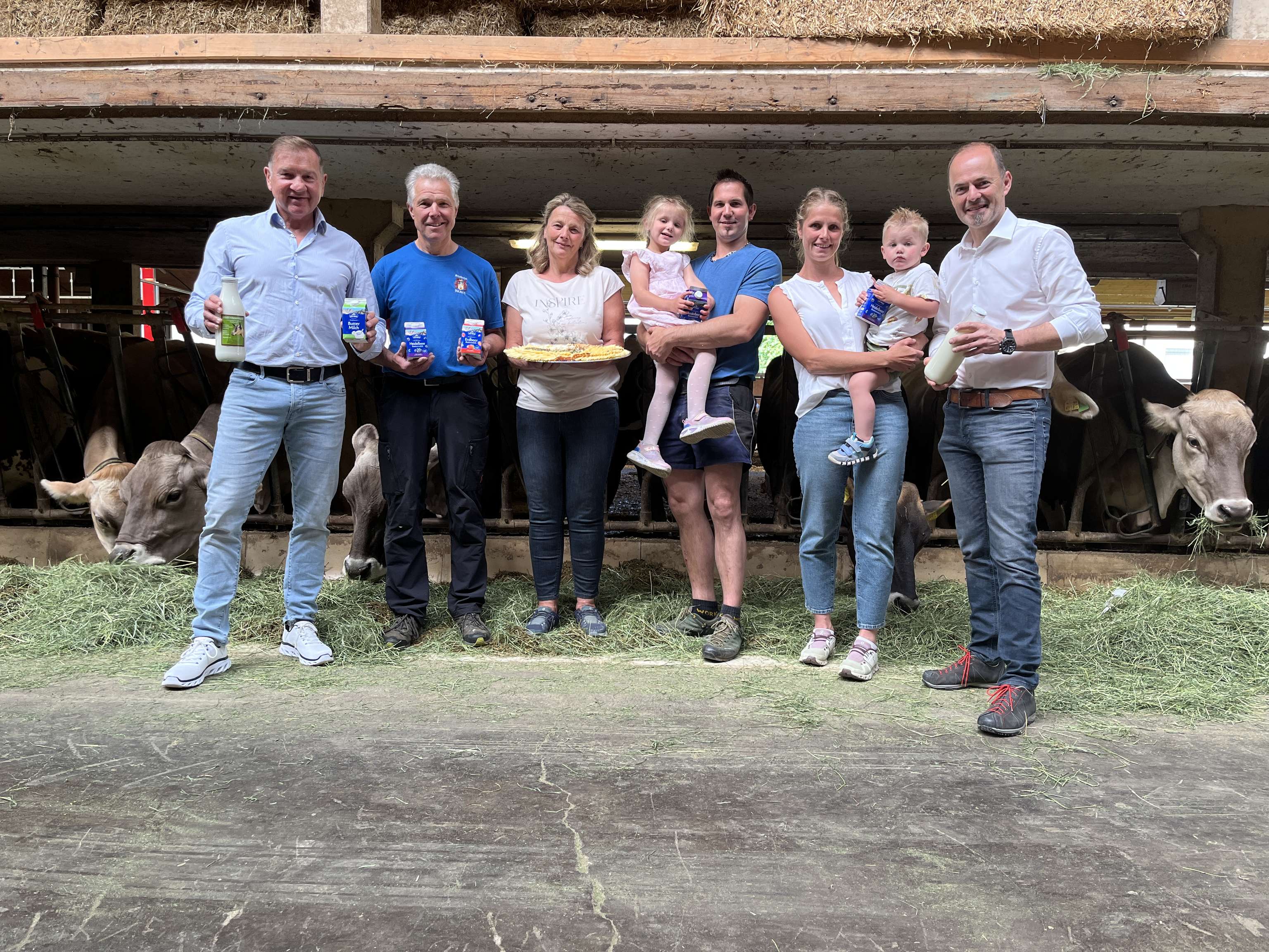 Eine Gruppe von Menschen steht in einem Kuhstall, hält Milchprodukte und ein Kind. Kühe fressen im Hintergrund.