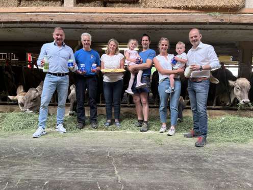 Eine Gruppe von Menschen steht in einem Kuhstall, hält Milchprodukte und ein Kind. Kühe fressen im Hintergrund.