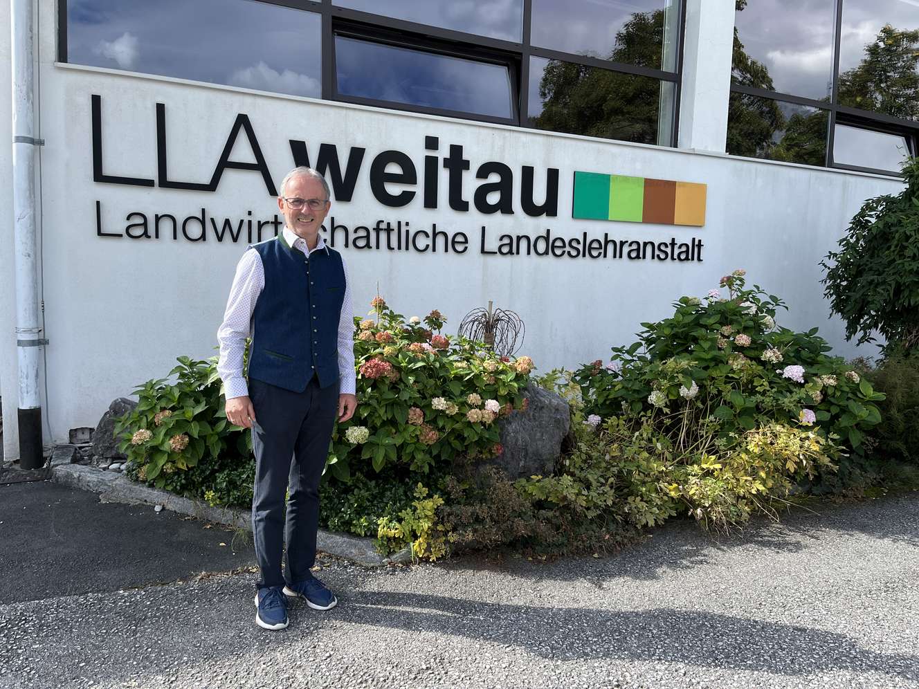 Mann mit Brille und blauer Weste steht vor dem Gebäude der LLA Weitau, Landwirtschaftliche Landeslehranstalt, umgeben von Hortensien.