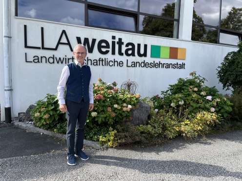 Mann mit Brille und blauer Weste steht vor dem Gebäude der LLA Weitau, Landwirtschaftliche Landeslehranstalt, umgeben von Hortensien.