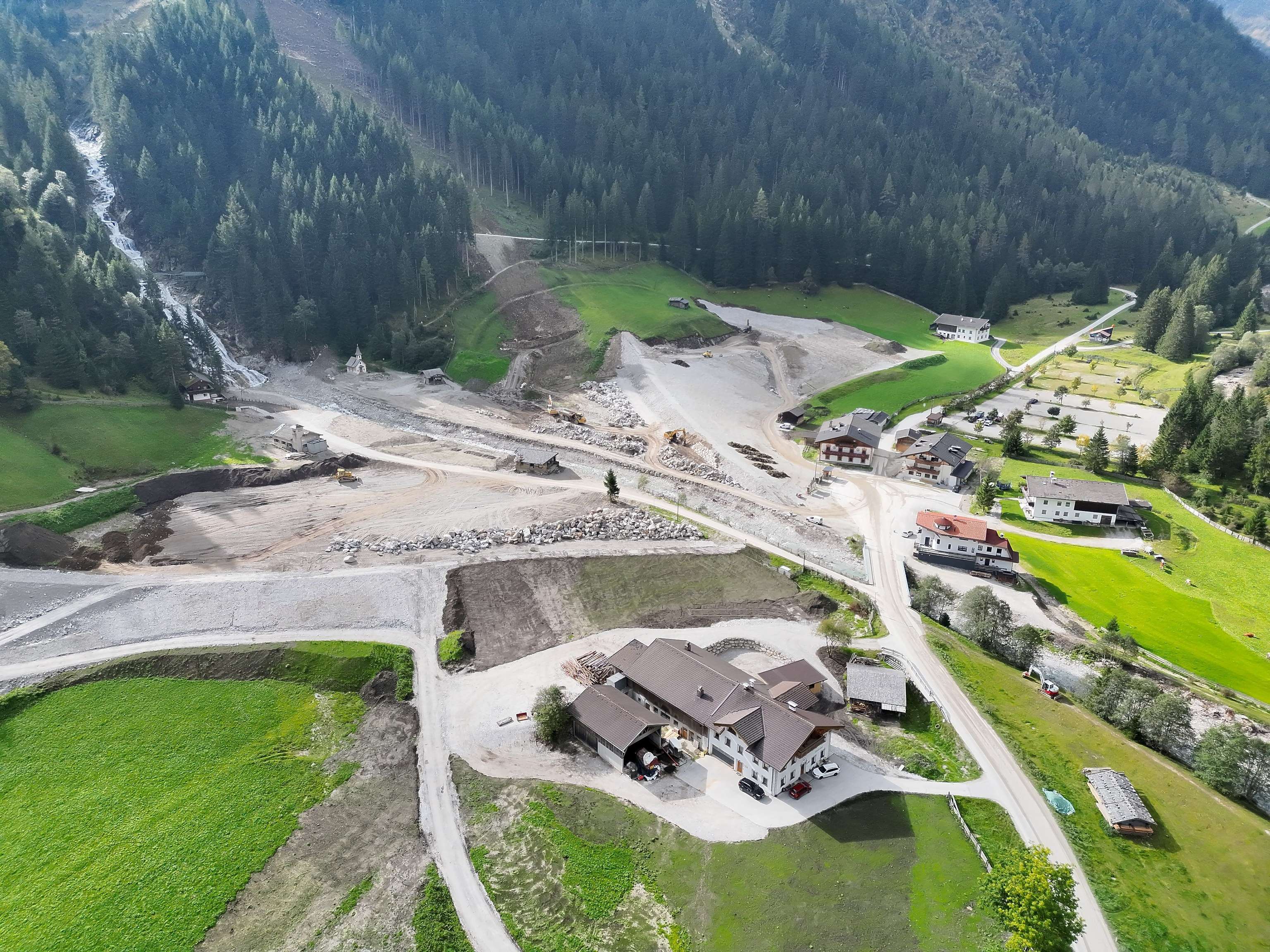 Luftaufnahme einer Baustelle in einem grünen, waldreichen Tal, mit mehreren Gebäuden und unbefestigten Straßen, umgeben von Bergen.