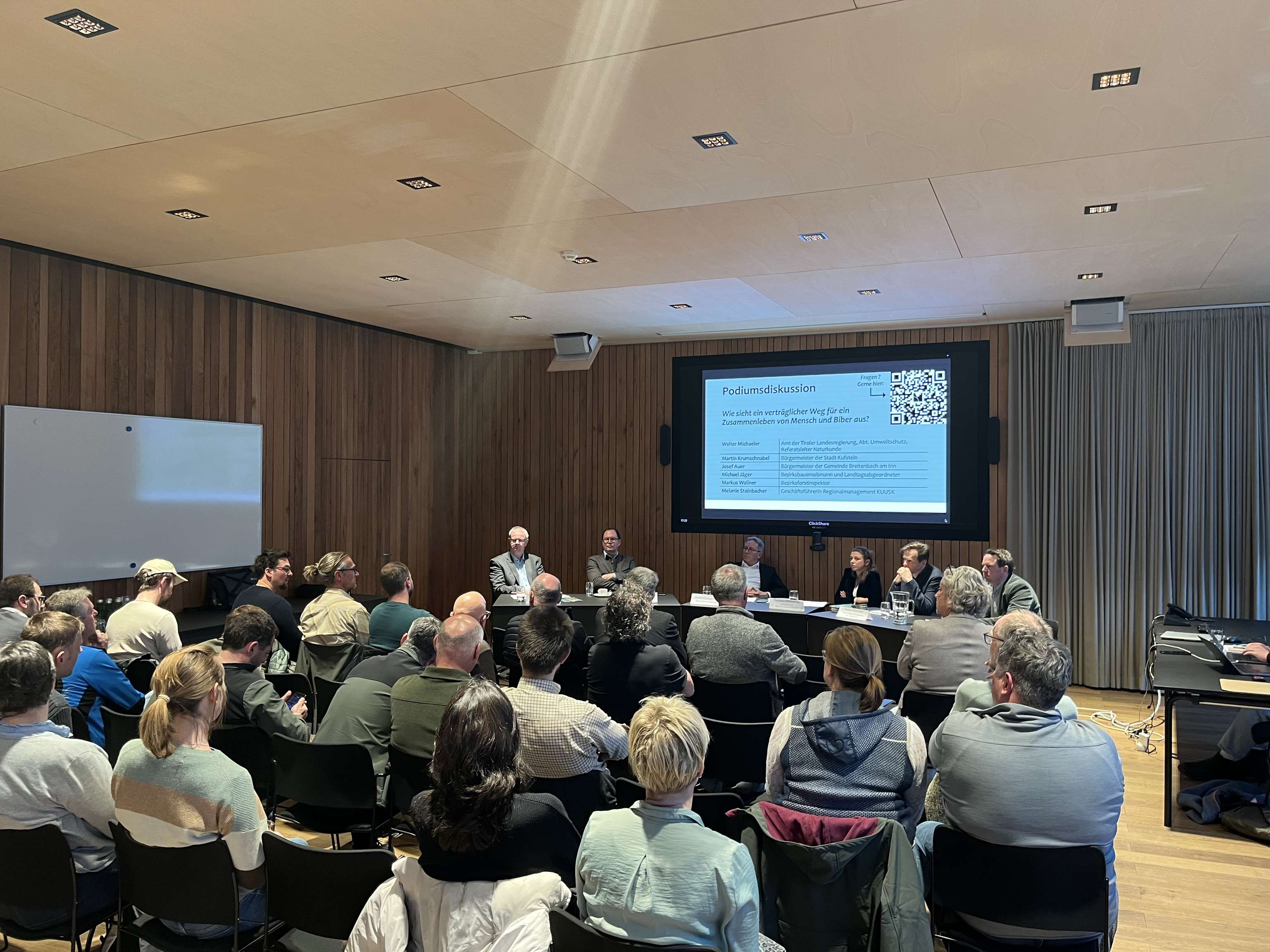 Podiumsdiskussion mit sieben Personen auf einer Bühne, Publikum sitzt in einem modernen Sitzungssaal, großer Bildschirm im Hintergrund.