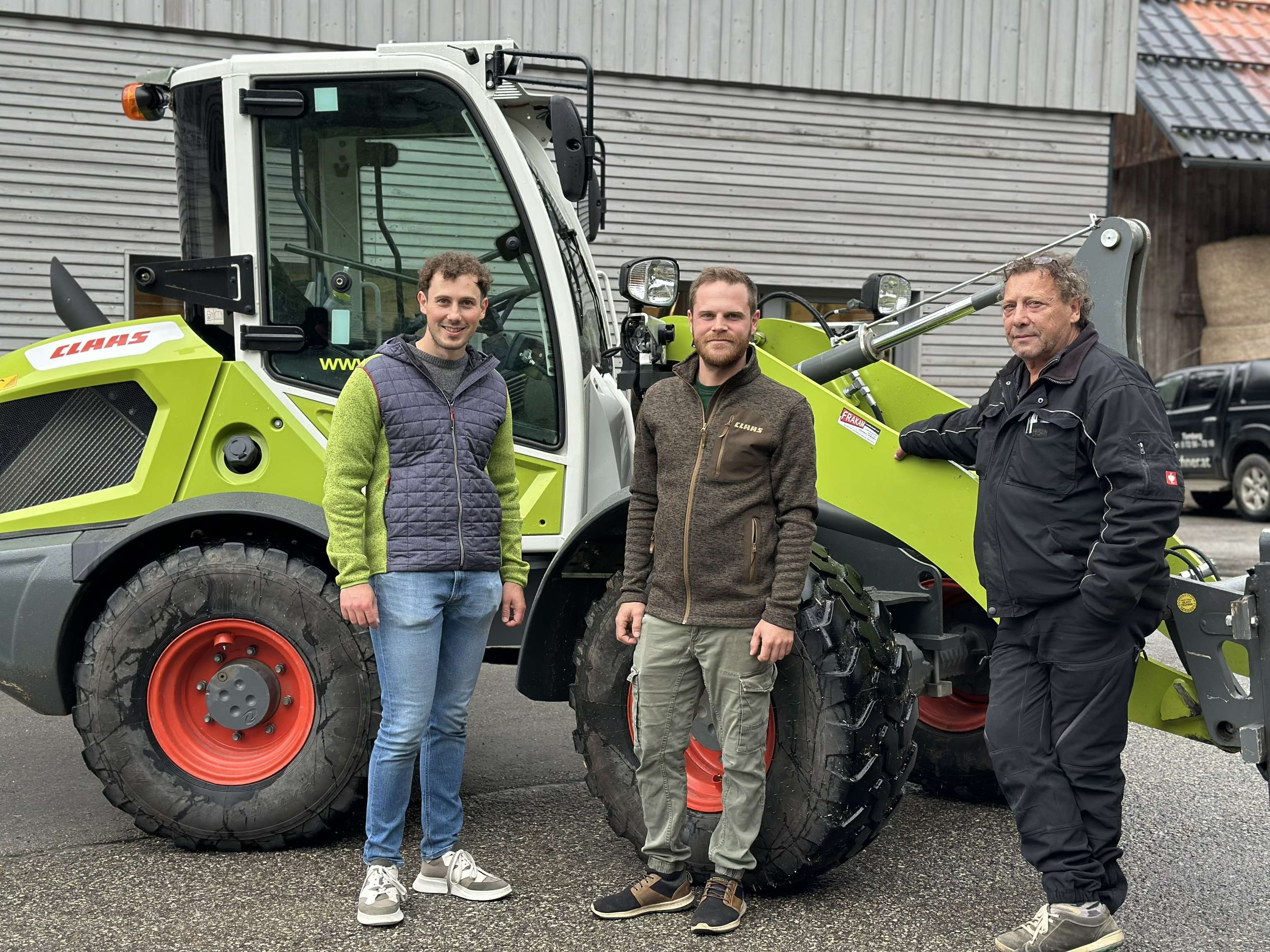 Drei Männer stehen vor einem Traktor. Sie tragen Freizeitkleidung und lächeln in die Kamera. Im Hintergrund sind Gebäude zu sehen.