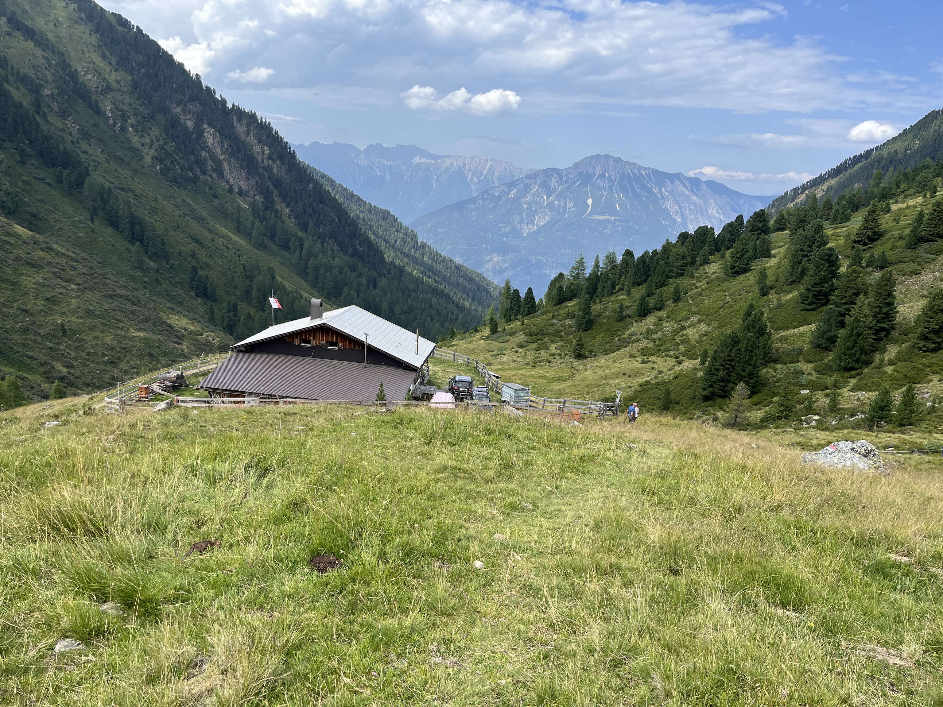 Alpine Berghütte auf grüner Almwiese mit italienischer Flagge, umgeben von Nadelwäldern und Bergpanorama unter blauem Himmel.