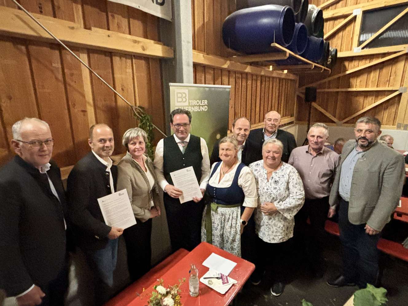 Gruppenfoto bei einer Veranstaltung des Tiroler Bauernbundes in einer Holzhalle, mehrere Personen halten Dokumente, teils in Tracht gekleidet.