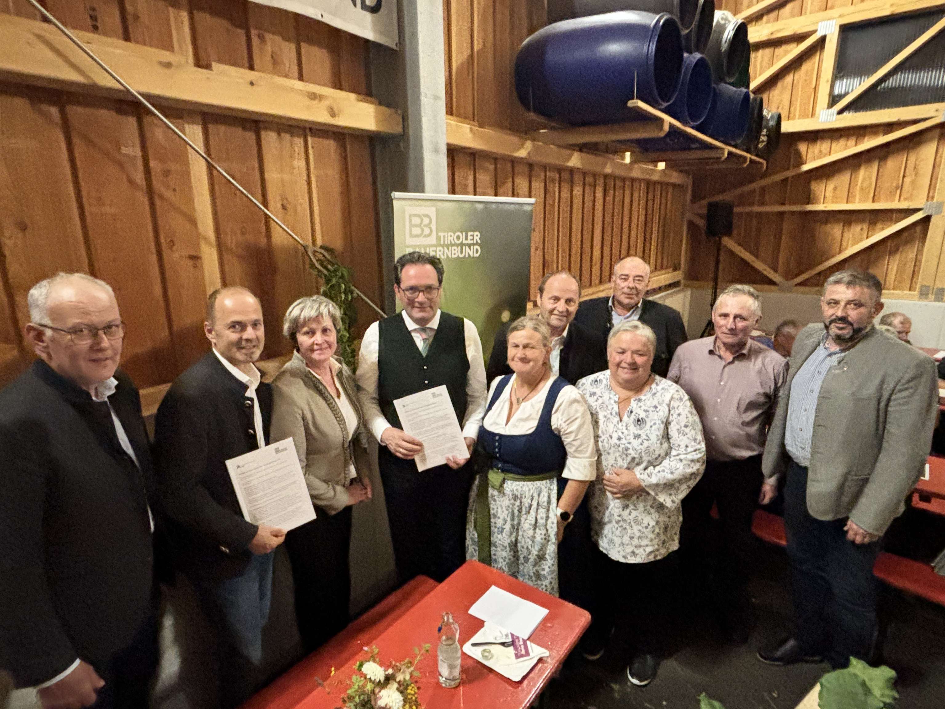 Gruppenfoto bei einer Veranstaltung des Tiroler Bauernbundes in einer Holzhalle, mehrere Personen halten Dokumente, teils in Tracht gekleidet.