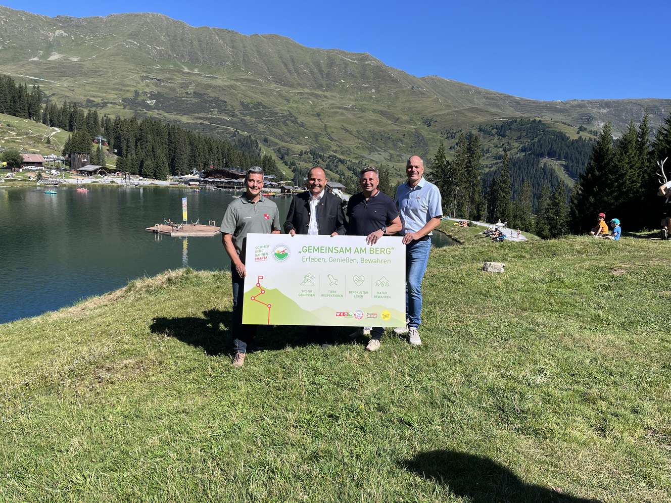 Vier Männer halten ein Schild mit der Aufschrift „Gemeinsam am Berg" vor einem Alpensee mit Bergen im Hintergrund.