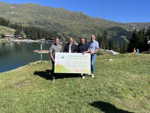 Vier Männer halten ein Schild mit der Aufschrift „Gemeinsam am Berg" vor einem Alpensee mit Bergen im Hintergrund.