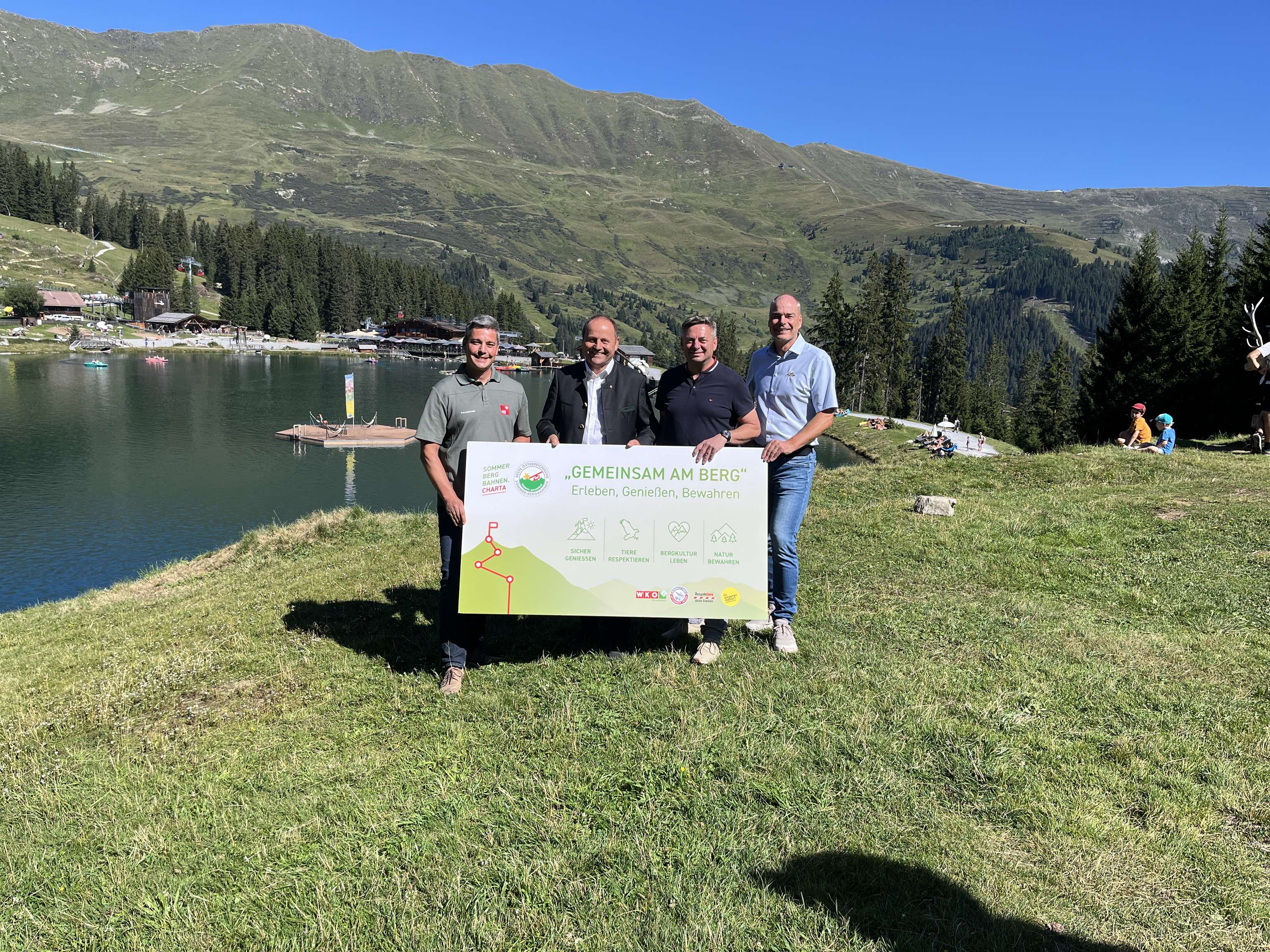 Vier Männer halten ein Schild mit der Aufschrift „Gemeinsam am Berg" vor einem Alpensee mit Bergen im Hintergrund.