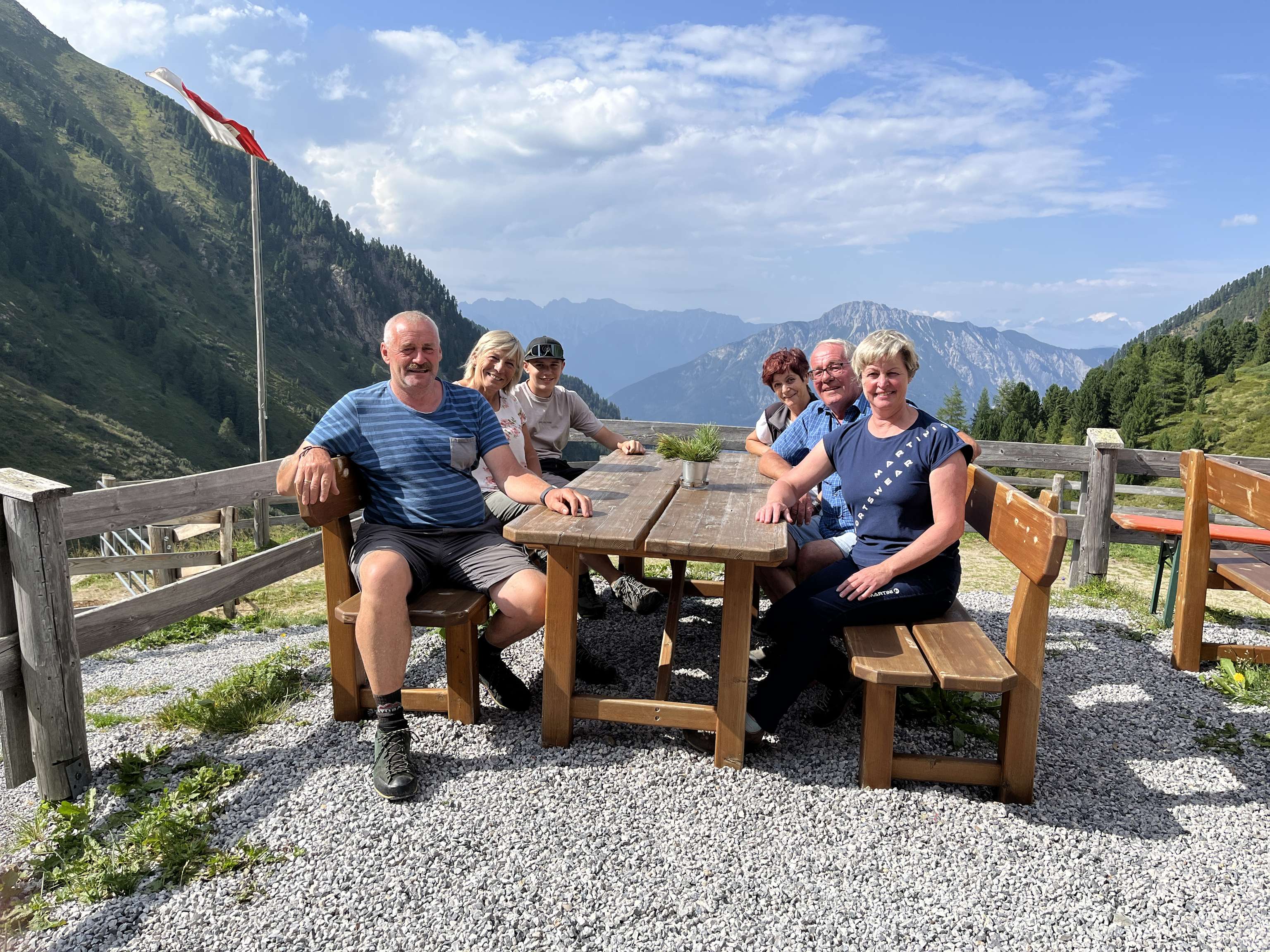 Gruppe von sechs Personen sitzt an einem Holztisch auf einer Almterrasse mit beeindruckendem Alpenpanorama und österreichischer Flagge.