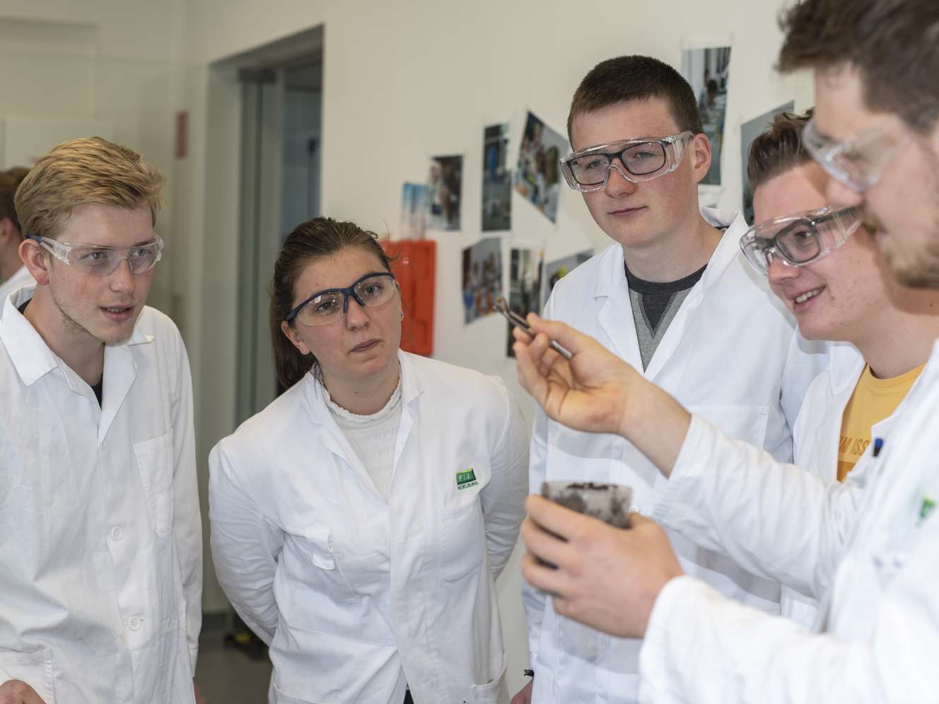 Vier Studierende in weißen Laborkitteln und Schutzbrillen betrachten ein Objekt im Labor.