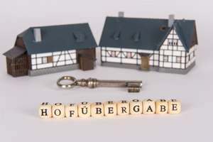 Zwei Spielzeug-Bauernhäuser mit einem Schlüssel und dem Schriftzug "Hofübergabe" davor.