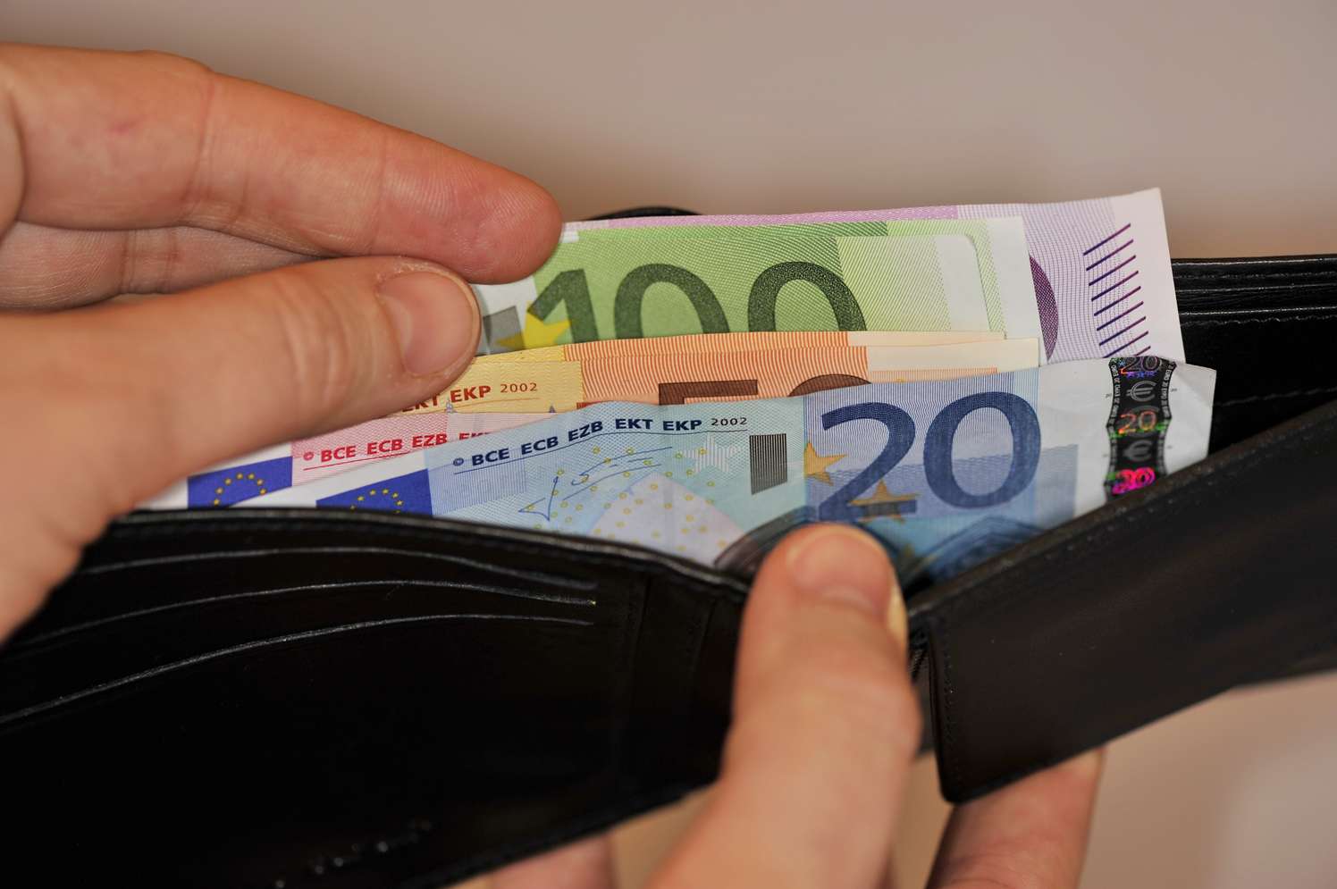 Hände halten ein schwarzes Lederportemonnaie mit mehreren Euroscheinen, darunter 100- und 20-Euro-Scheine.