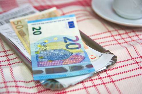 Euroscheine (20 und 50 Euro) mit Kassenbon auf einem Zahlungstablett im Restaurant, rot-weißes Tischtuch.