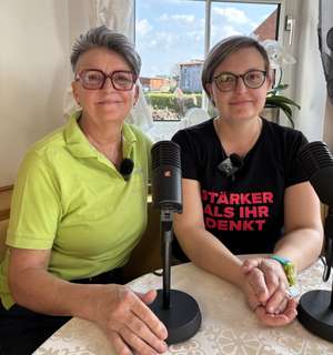 Zwei Frauen mit Brillen sitzen an einem Tisch mit einem Podcast-Mikrofon. Eine trägt ein schwarzes Shirt mit „Stärker als ihr denkt".