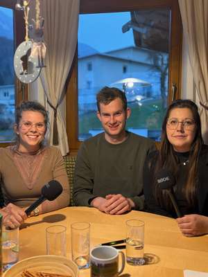 Drei Personen sitzen am Tisch mit Mikrofonen, lächeln und schauen in die Kamera. Hintergrund zeigt Fenster mit Blick nach draußen.