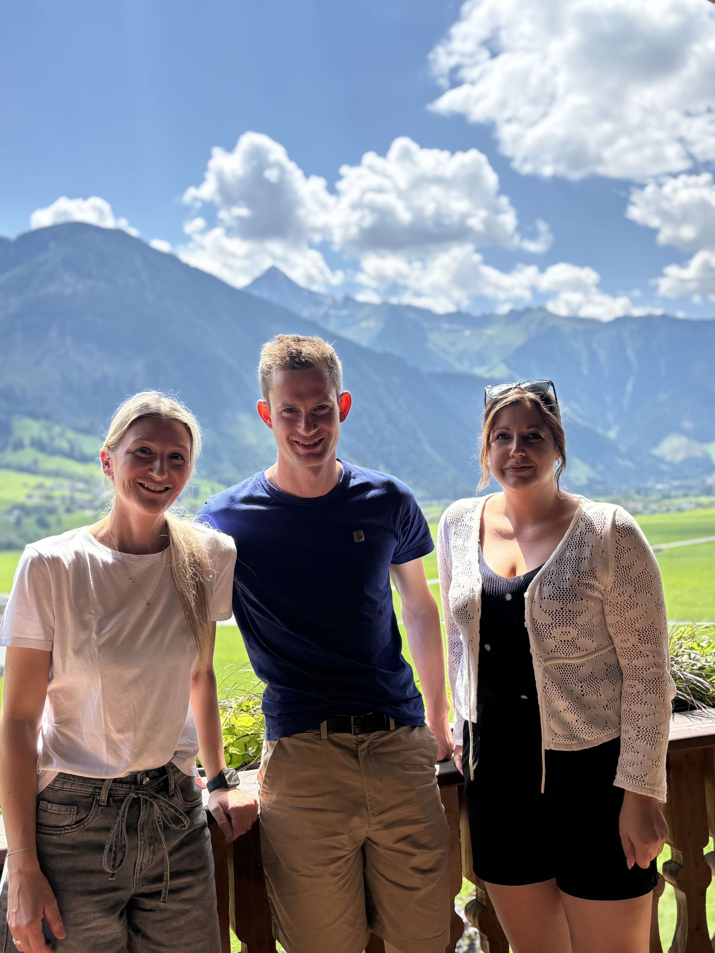 Drei lächelnde Personen auf einer Terrasse mit grüner Almlandschaft und Bergen im Hintergrund bei sonnigem Wetter.