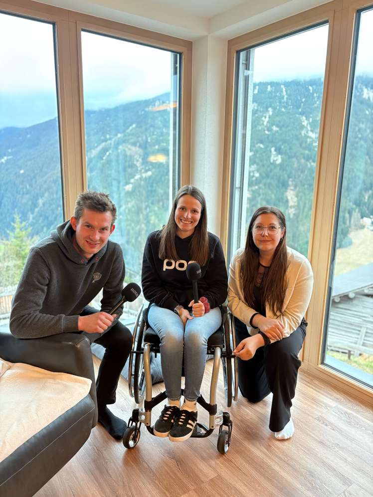 Drei Personen mit Mikrofonen sitzen vor großen Fenstern mit Bergblick. Eine Person sitzt im Rollstuhl.