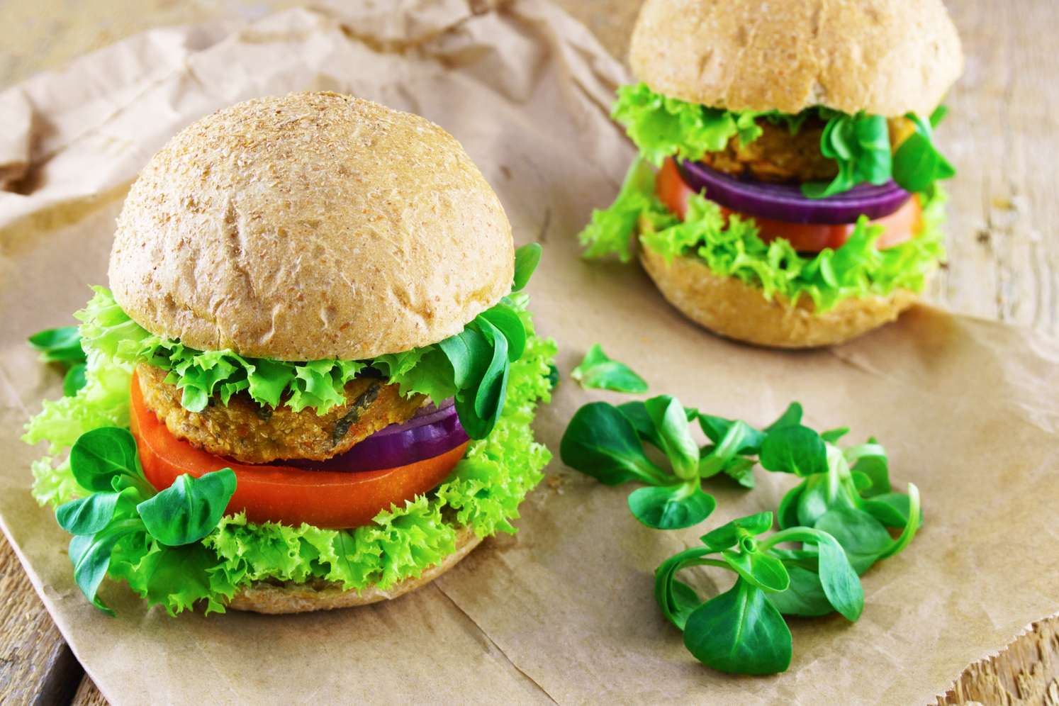 Zwei vegane Burger mit Vollkornbrötchen, Salat, Tomate, roter Zwiebel und pflanzlichem Patty auf Backpapier.