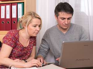 Frau und Mann schauen konzentriert auf einen Laptop im Büro, im Hintergrund rote Ordner mit Beschriftungen wie Buchhaltung und Bilanzen.