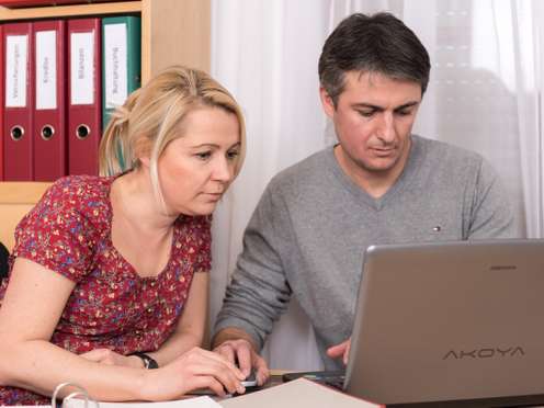 Frau und Mann schauen konzentriert auf einen Laptop im Büro, im Hintergrund rote Ordner mit Beschriftungen wie Buchhaltung und Bilanzen.