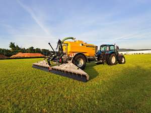 Blauer Traktor mit gelbem Zunhammer Güllefass 8000 und Schleppschuhverteiler auf grüner Wiese bei sonnigem Wetter.