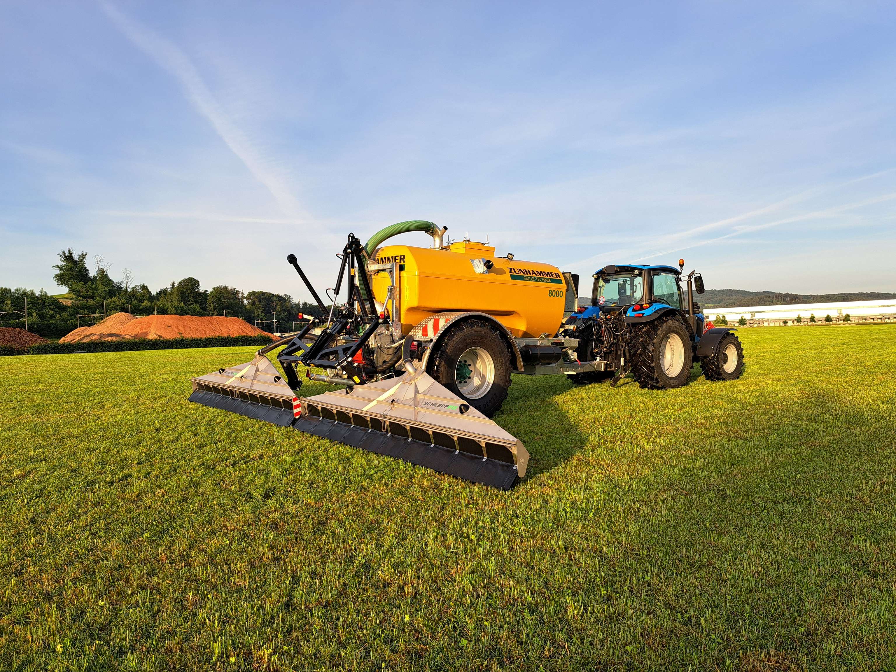 Blauer Traktor mit gelbem Zunhammer Güllefass 8000 und Schleppschuhverteiler auf grüner Wiese bei sonnigem Wetter.