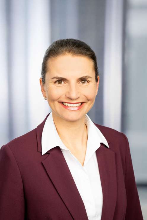 Lächelnde Frau im bordeauxroten Blazer mit weißem Hemd, hochgesteckten Haaren, professionelles Porträtfoto vor unscharfem Hintergrund.