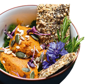 Schale mit orangener Creme, garniert mit Samen, essbaren Blumen, Kräutern und knusprigem Knäckebrot.