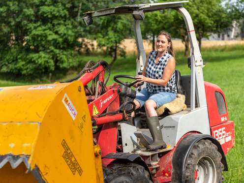 Junge Frau fährt einen roten Weidemann Hoflader mit gelbem Anbaugerät auf einer grünen Wiese im Sommer.