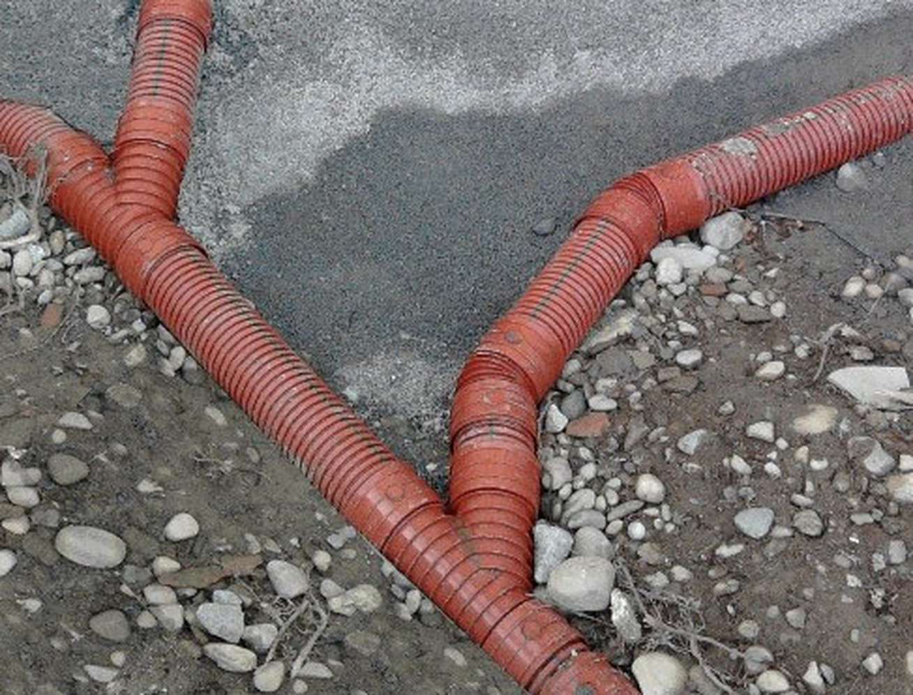 Rote gewellte Kunststoffrohre auf kiesigem Untergrund, die sich verzweigen und auf einer Baustelle verlegt sind.