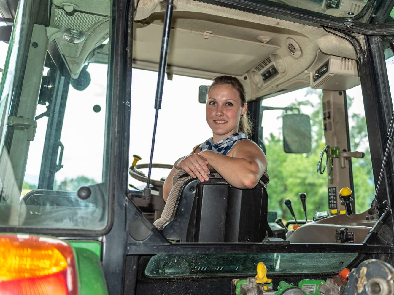 Lächelnde junge Frau sitzt in der Fahrerkabine eines grünen Traktors und stützt sich auf den Sitz.