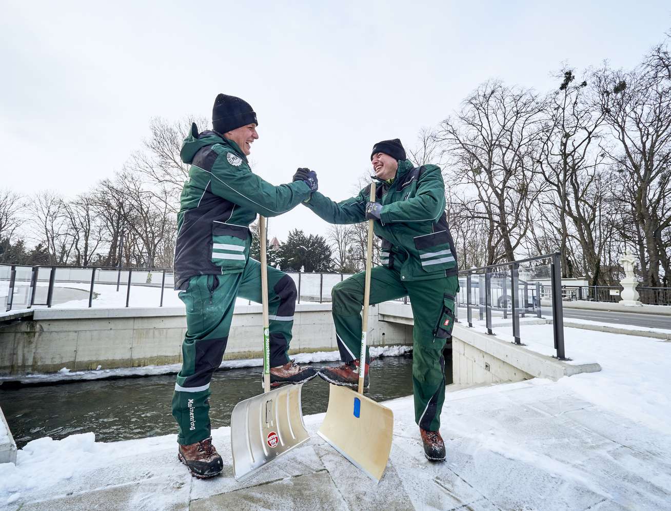 Zwei lachende Mitarbeiter in grünen Arbeitsanzügen geben sich die Hand, mit Schneeschaufeln im winterlichen Park.