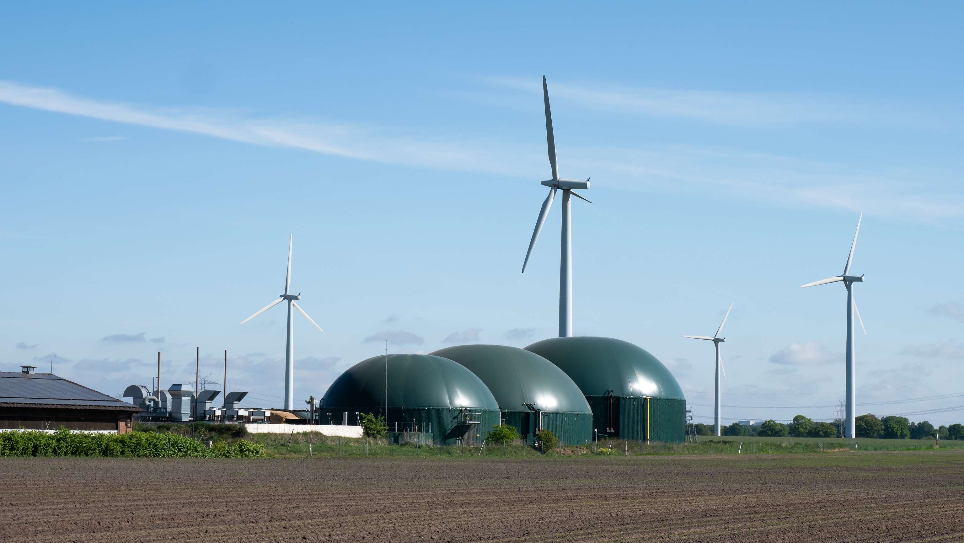 Drei Bioreaktoren auf einer Freifläche, dahinter Windräder