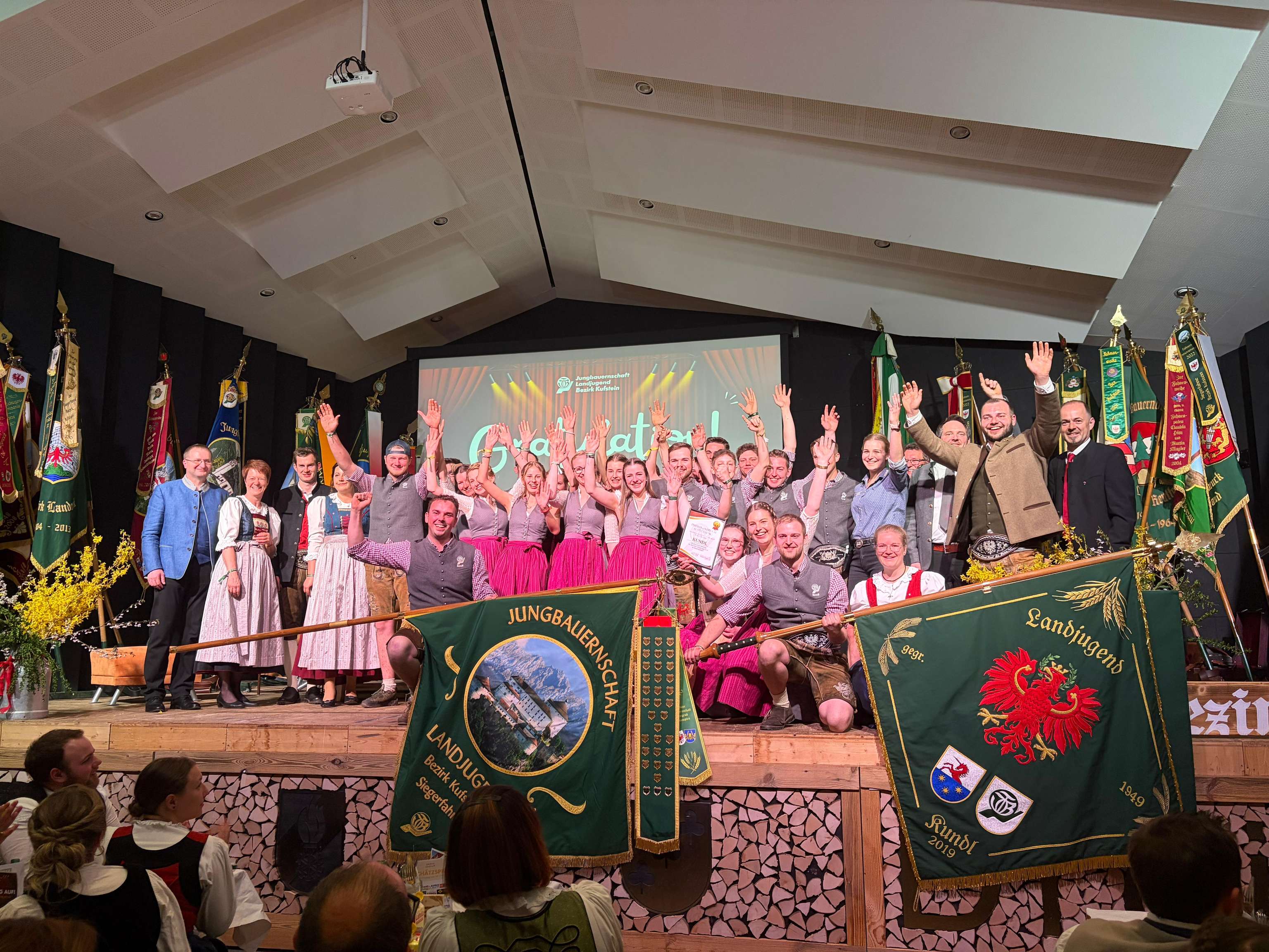 Eine Gruppe in traditioneller Tracht steht auf einer Bühne und hält Fahnen, während sie in die Kamera lächelt und die Arme hebt.