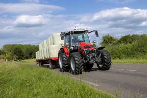 A red tractor transports large wrapped hay bales on a trailer along a rural road, surrounded by greenery and under a partly cloudy sky.