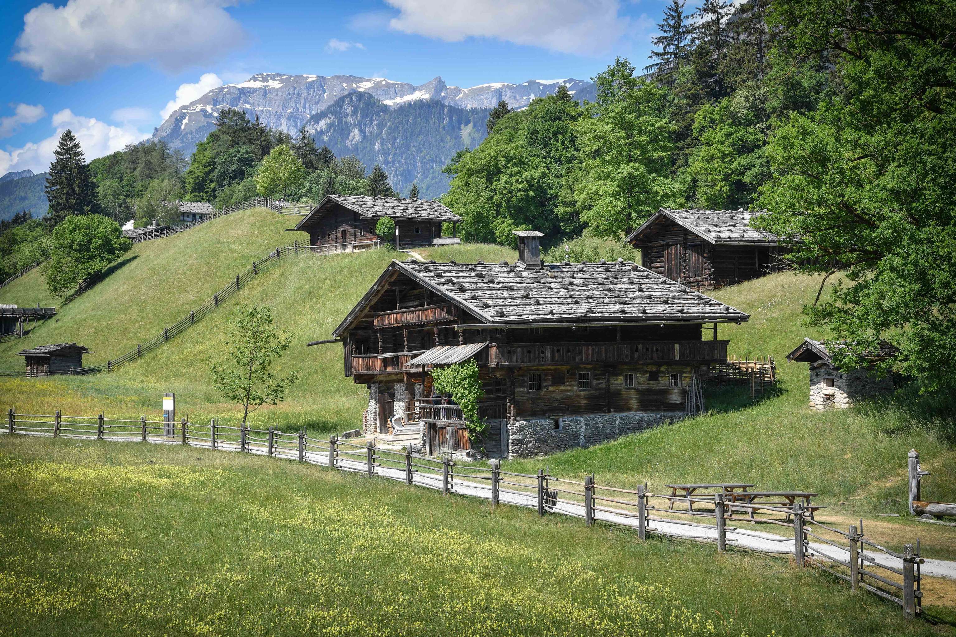 Alpenlandschaft mit traditionellen Holzhäusern auf grünen Wiesen, Bergblick im Hintergrund, blauer Himmel mit Wolken.
