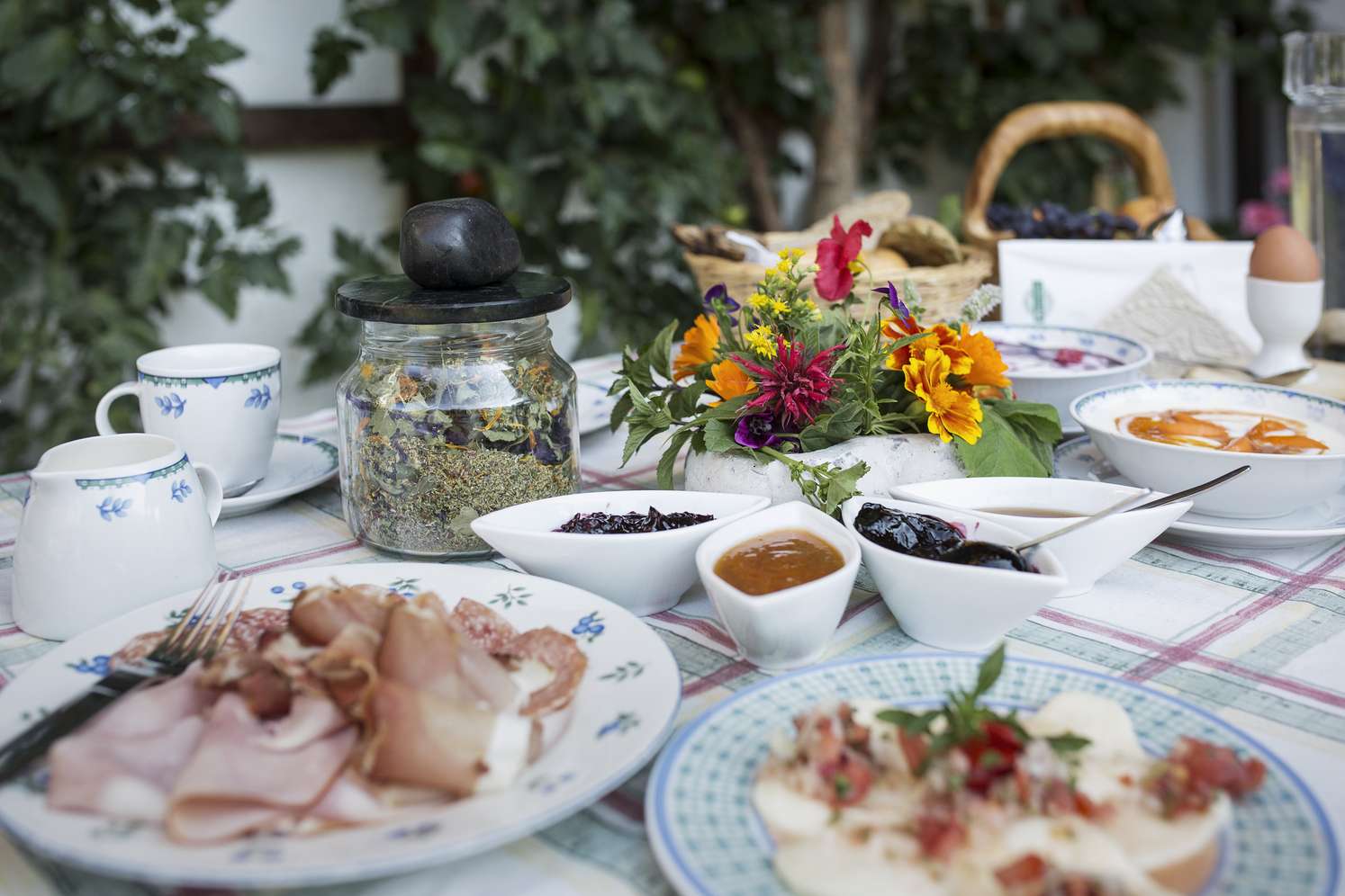 Ein gedeckter Tisch mit Aufschnitt, Dips, Butter, Blumenarrangement und Kräuterglas vor grünem Hintergrund.