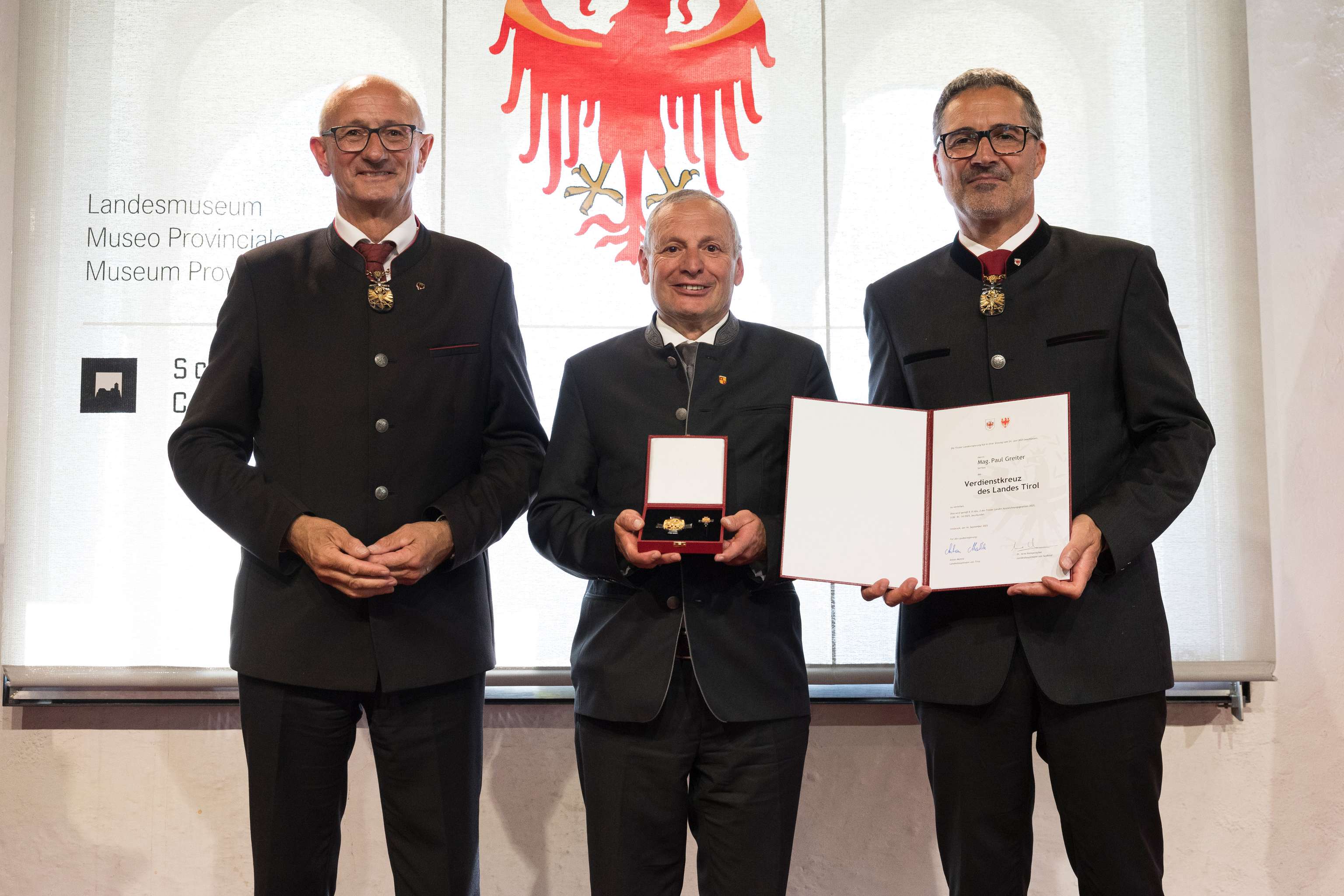 Three men in formal attire stand indoors; the center man holds a medal and certificate, with a red eagle emblem in the background.